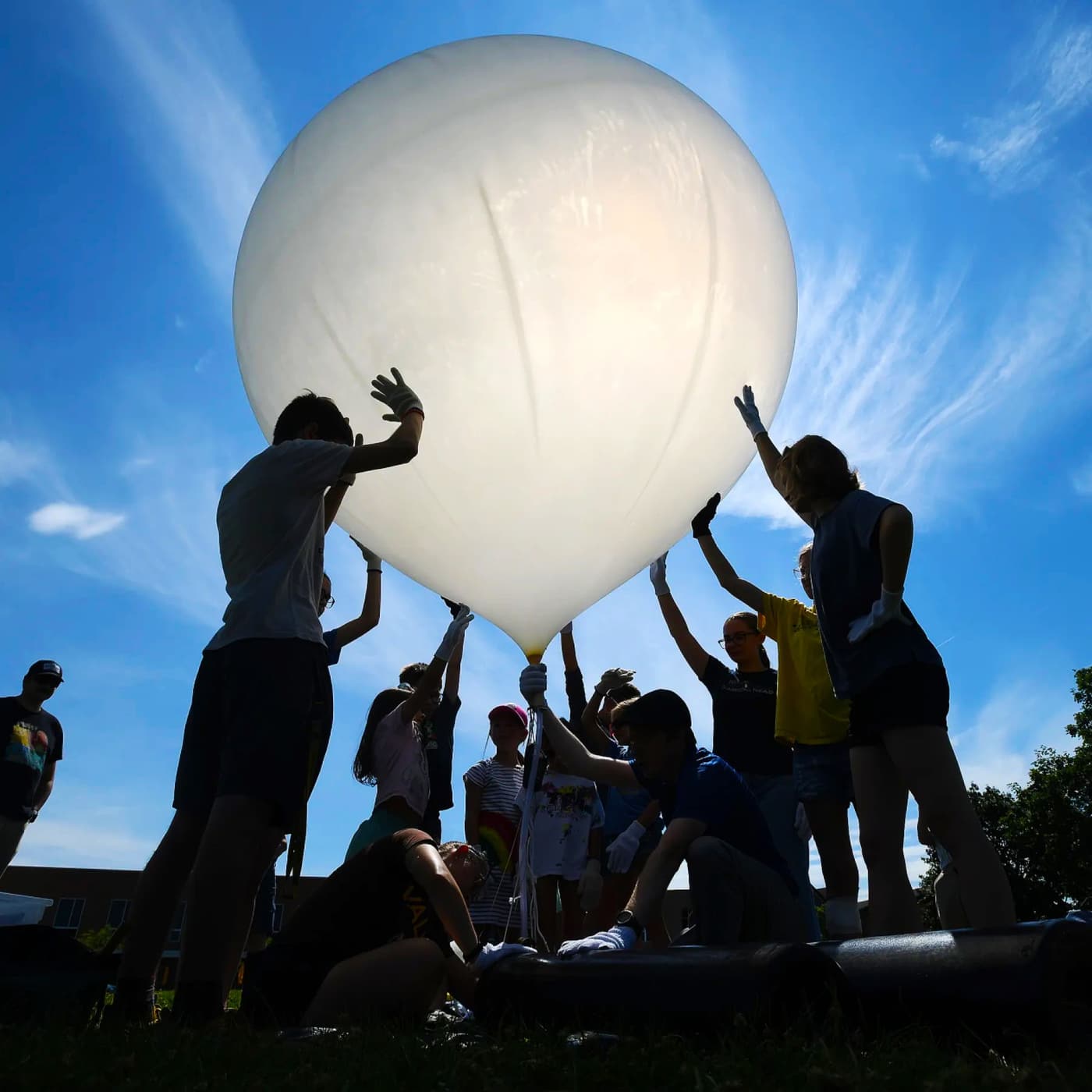 A natural latex weather balloon lifting off from a coastal field, with the payload string held by the launch crew.
