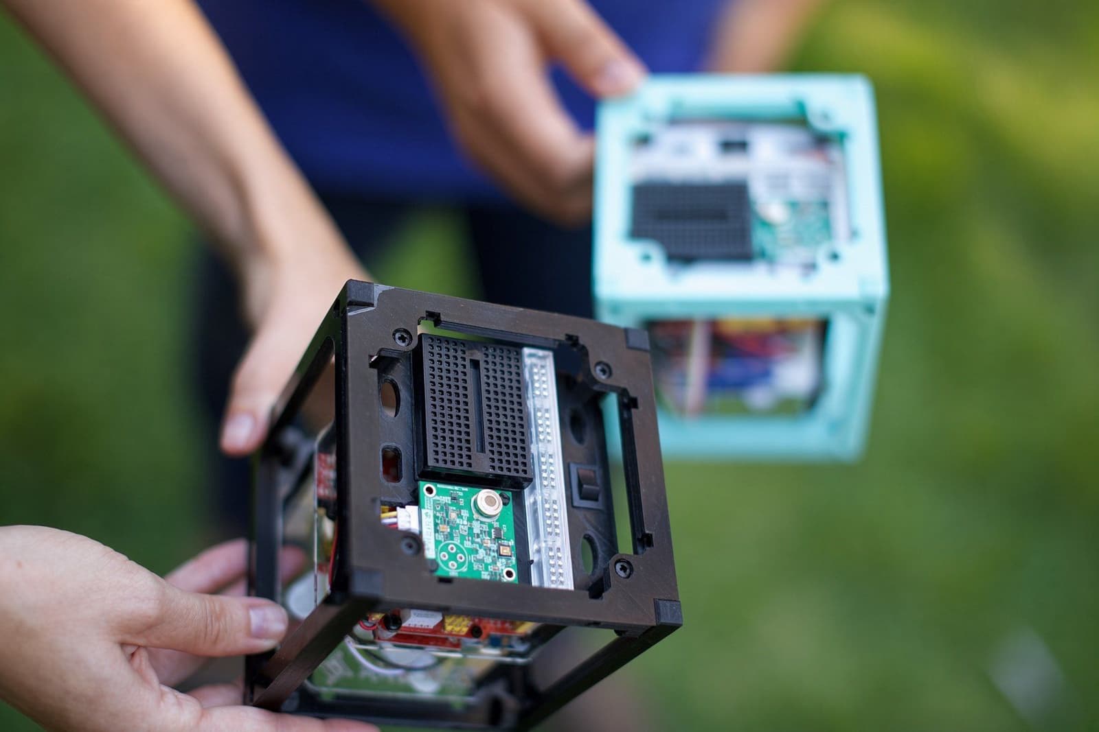 Students holding 3D-printed 1U CubeSats outdoors, with the open frame showing stacked flight boards.
